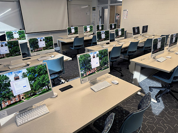 An image depicting a room with 6 tables, each table having 4 computer setups. The tables are set in two collumns side by side.